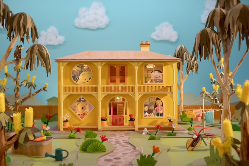 A paper-look two-storey yellow house sits on a landscaped block of land with Play School characters visible in the windows