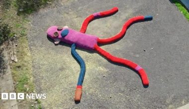 A sculpture of a giant sock puppet lying in a car park. It has a pink body, one blue and one red arm and two red legs. It has two giant button shapes for eyes.