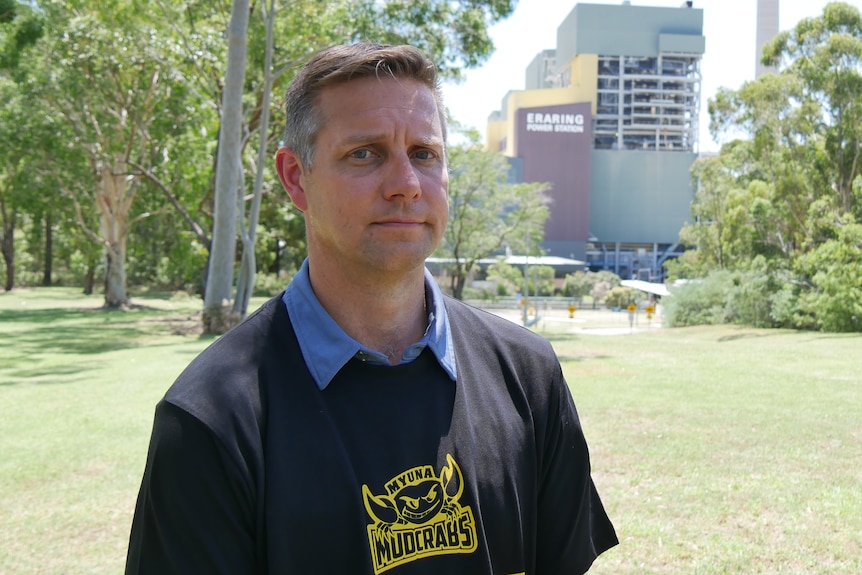 A man wearing a black t-shirt stands on grass in front of a power station. 
