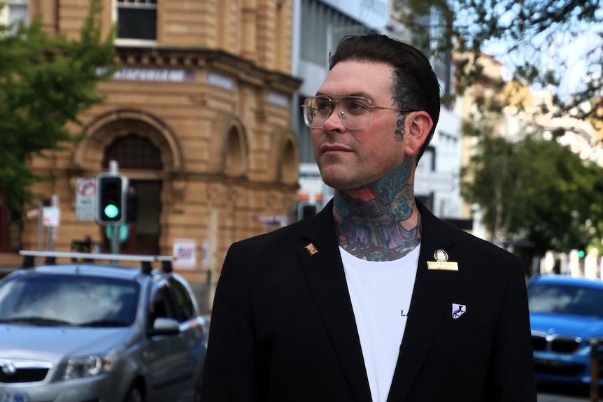 A heavily tattooed man wearing a white shirt and a black blazer stands on a busy street corner.
