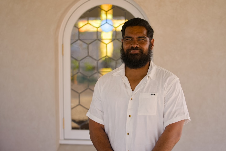 A bearded man in a white shirt stands in front of a chapel window with a stained-glass cross visible behind him.