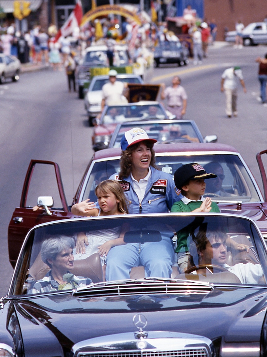 Wearing blue overalls and cap, a woman sits in an open-top car on parade, with one arm around her two children.