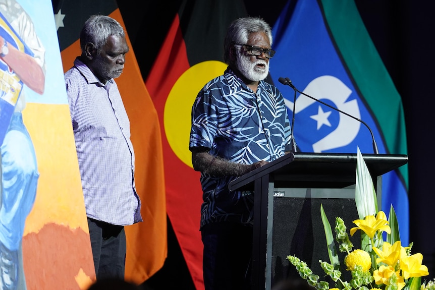 An Aboriginal man standing at a podium, gray hair and beard, blue pattern button up shirt, flags behind him.