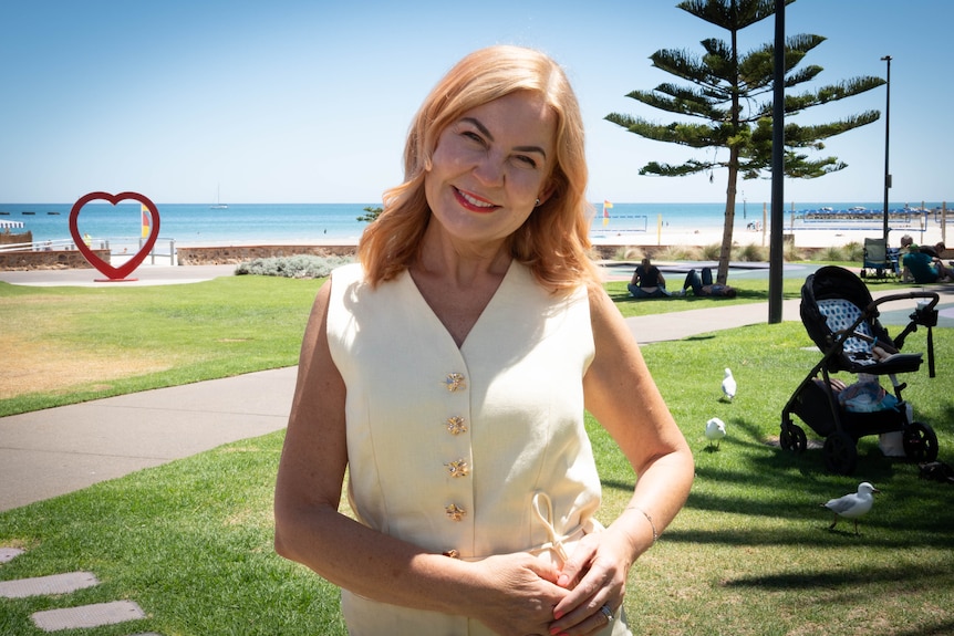 A blonde woman wearing a white sleeveless top standing on lawn near the beach with a love-heart sculpture