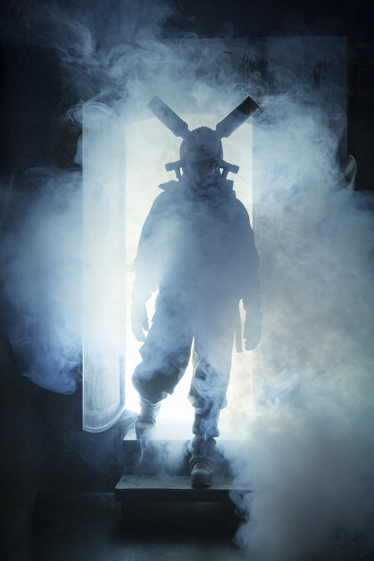 A smoke-hazed silhouette of a person in a steampunk outfit with mask, gloves and boots steps through a brightly lit door.