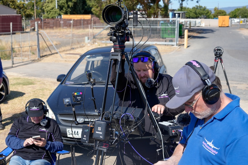 People with camera equipment in front of a black car