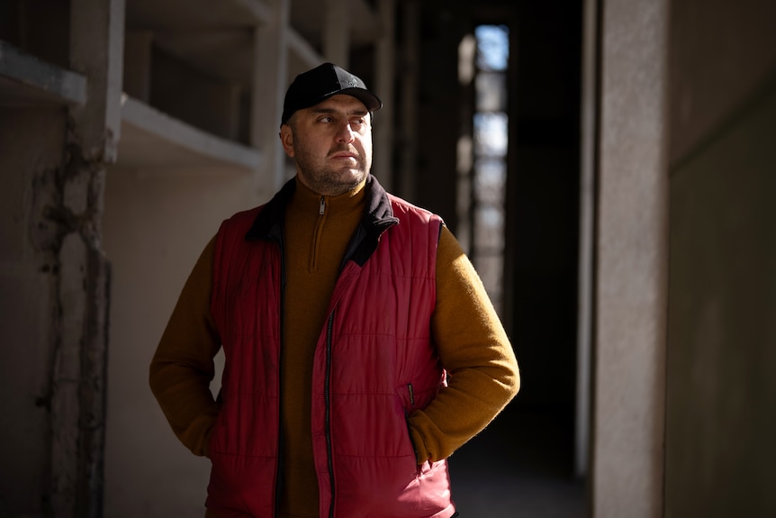 A man wearing a cap stands in a darkened industrial room. He is looking to the side with a serious expression.