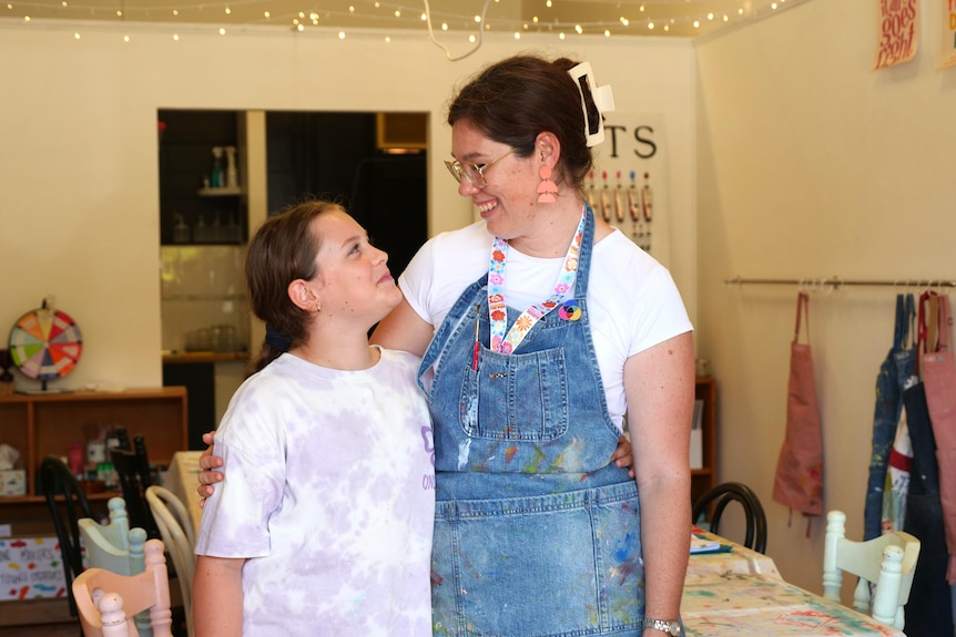 Deanna wearing an art apron with her arm around her daughter smiling down at her. 