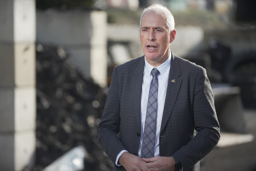 A man with short white hair wearing a suit and tie stands outside a concrete building.