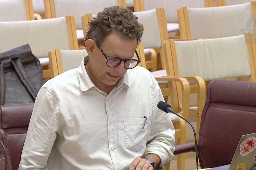 A man with curly brown hair, glasses and a crumpled shirt checks his notes. 
