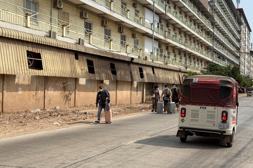 People drag luggage down a dusty street next to a multistory compound.
