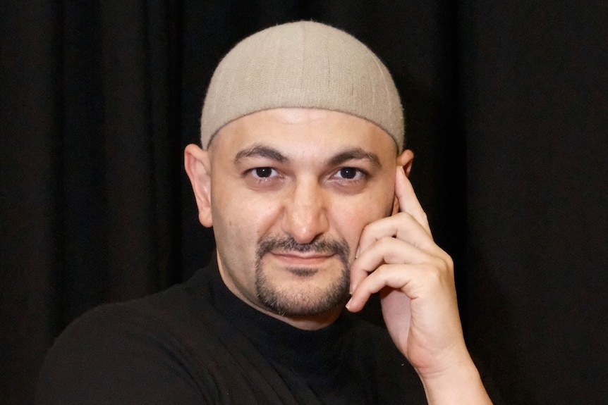 A Lebanese Australian man wearing a tan skull cap and a black t-shirt, standing against a black background.