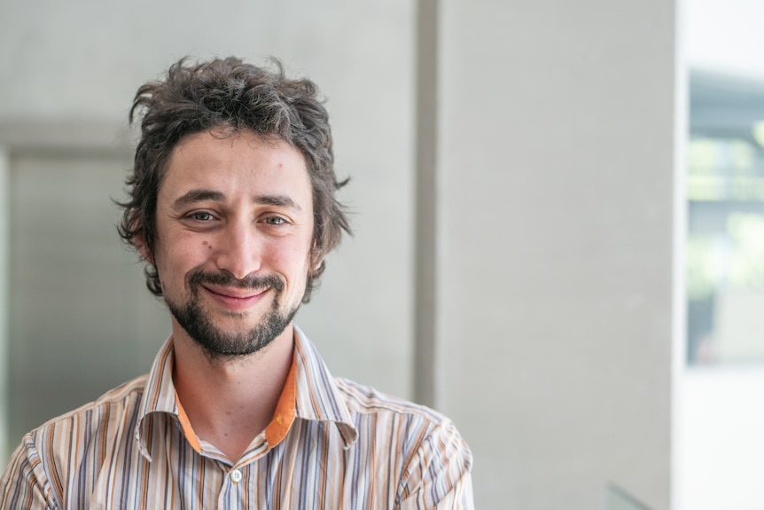 Young man with dark hair, beard, striped shirt smiles at camera.