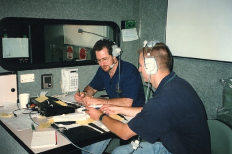 Two individuals wearing headsets sit at a desk covered in papers and equipment.