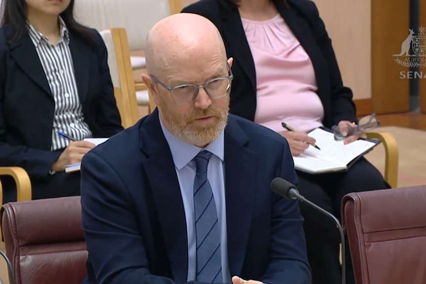A bearded man with glasses in a blue suit speaks into a microphone