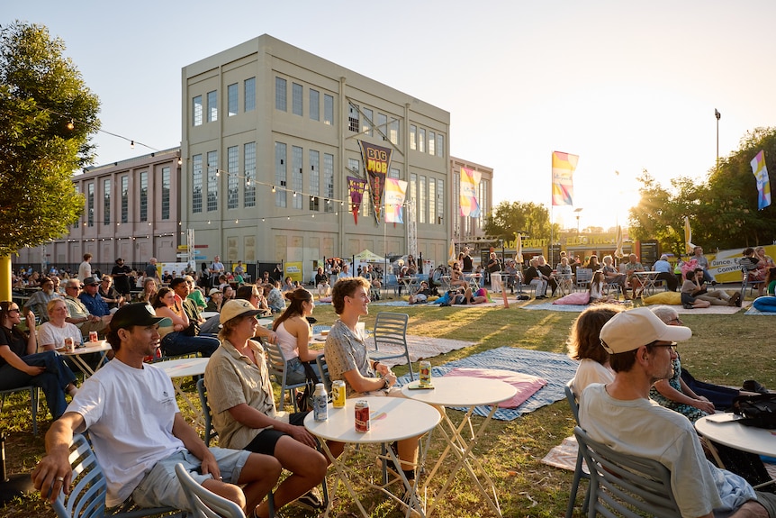 People sitting at tables and chairs on lawn with large building in background