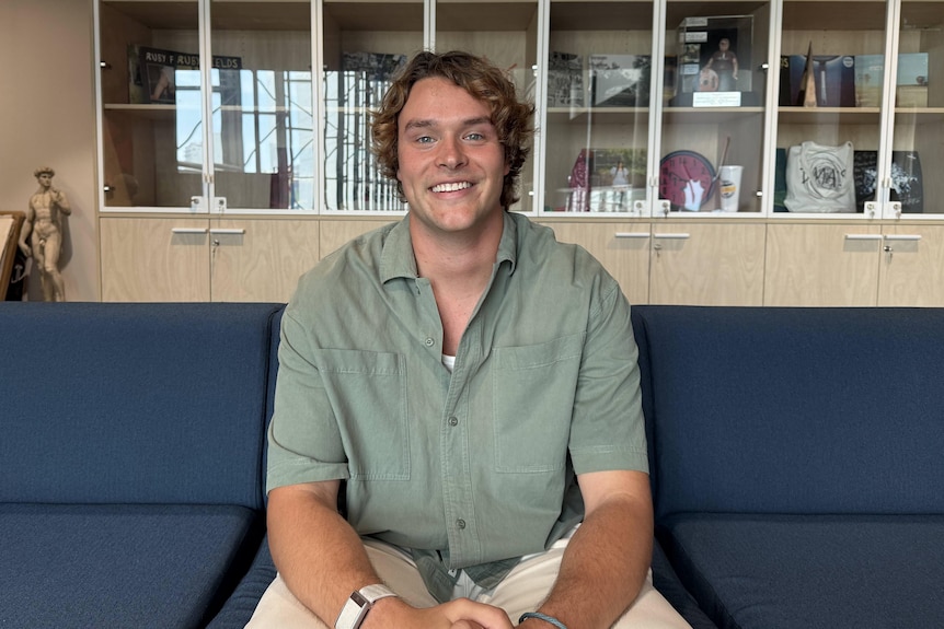 A white man with blonde hair sits, smiling, looking at camera in a green short sleeve shirt