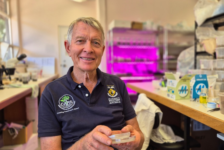 A man with glasses sits in a university lab and smiles at the camera