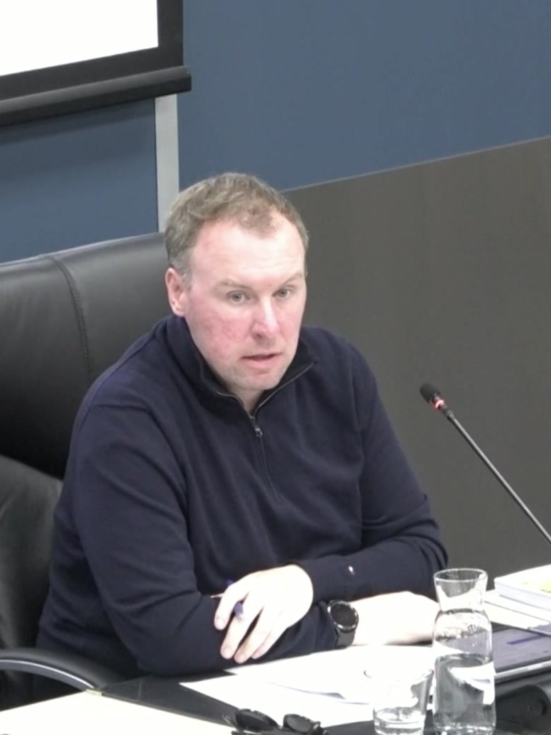 A man with sandy coloured hair in a blue top sits at a desk behind a microphone.