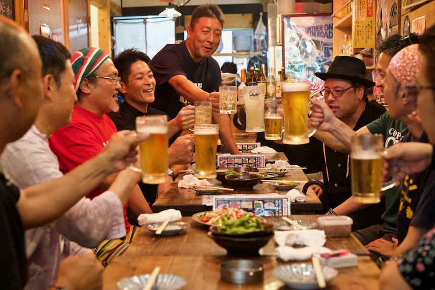 A group of smiling men seated around a table raise their glasses of beer in a toast.