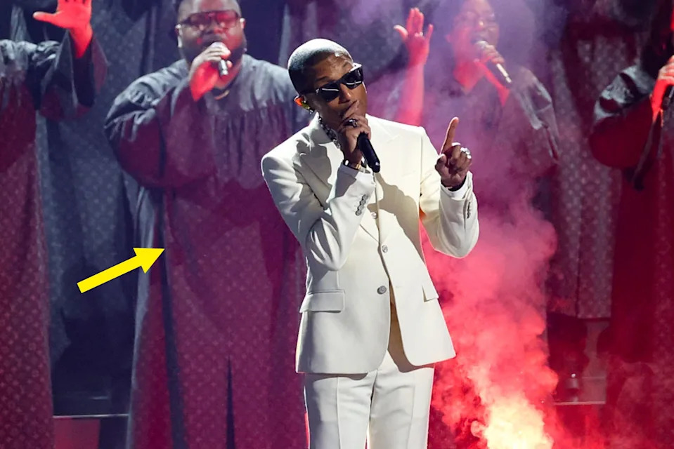 Person in a white suit sings passionately on stage, backed by a choir in robes, with dramatic stage smoke