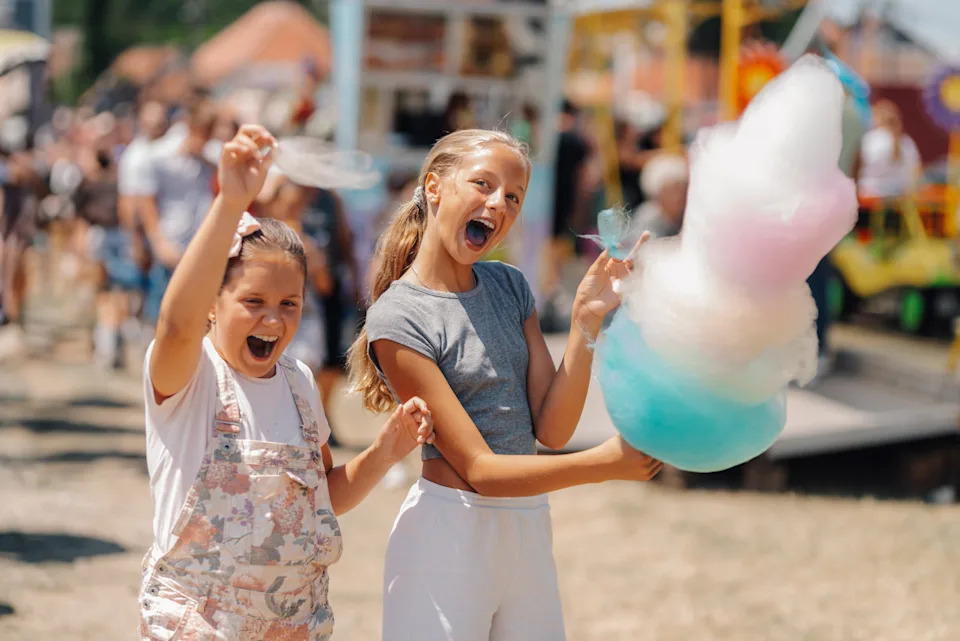 Two excited girls at a fair holding cotton candy, celebrating and enjoying a sunny day at an outdoor event