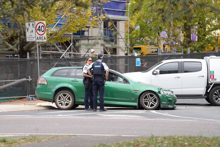 Two police officers stand talking seriously in front of a crashed green Holden Commodore.