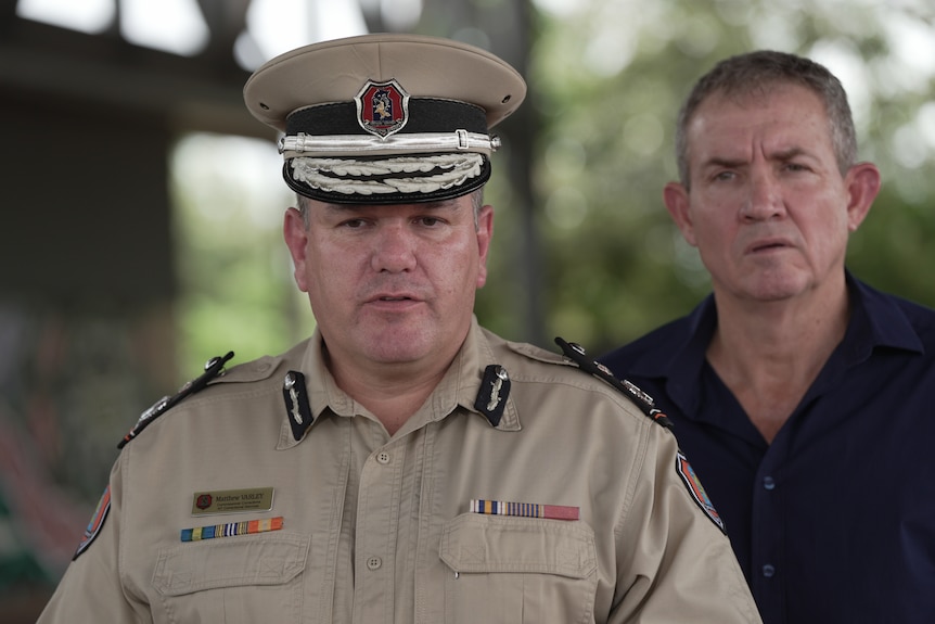 A white man in a beige uniform, with maroom emblem on his cap, staring at camera. Man in navy blue button down behind him