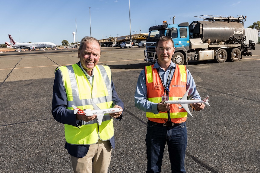 Two men in high-vis workwear holding miniature model aeroplane on the tarmac at an airport.