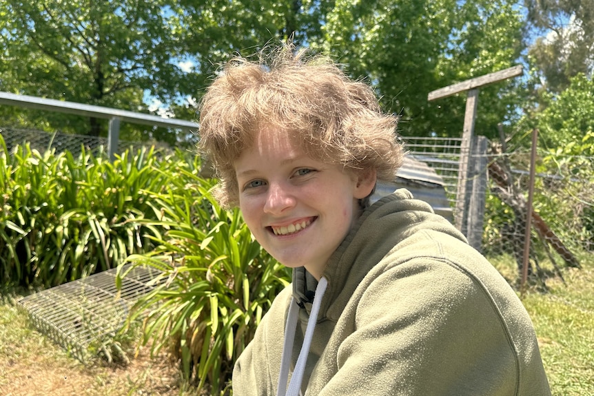 A teenager with short blonde hair smiles at the camera while sitting on green grass in front of trees and shrubs.