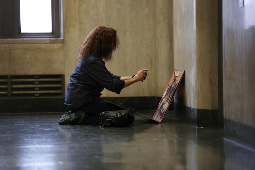A woman on the ground takes a photo of a painting. 