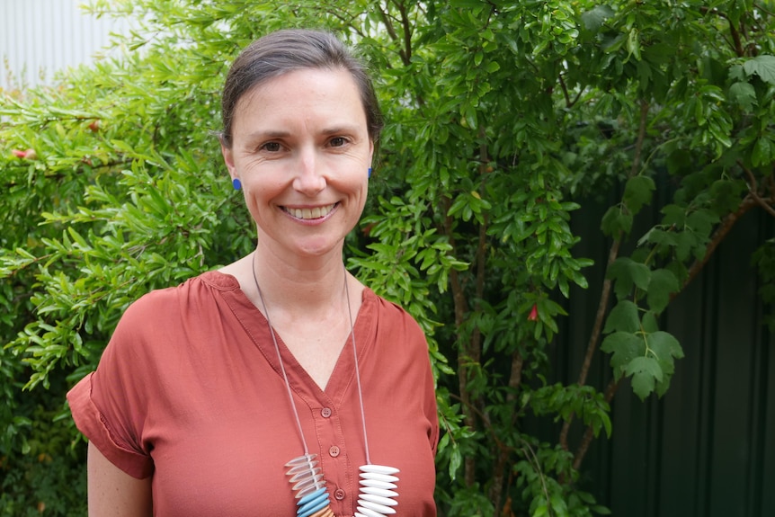 Olivia in an orange top smiling at the camera, stands in front of a bush or tree with red flowers.
