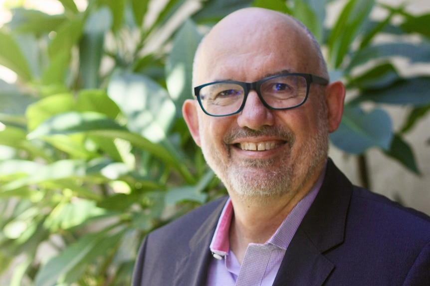 Portrait of a bald man with grey stubble beard, dark rimmed glasses and a navy suit jacket in front of a plant.
