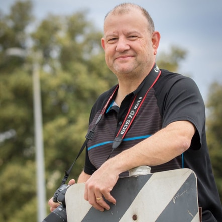 A man with a camera strap around his neck leaning on a sign