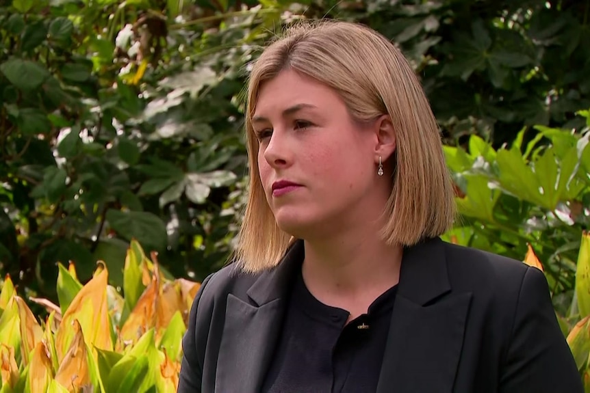 A woman with shoulder length blonde hair in a black jacket and top stands near bushes, looking off camera.