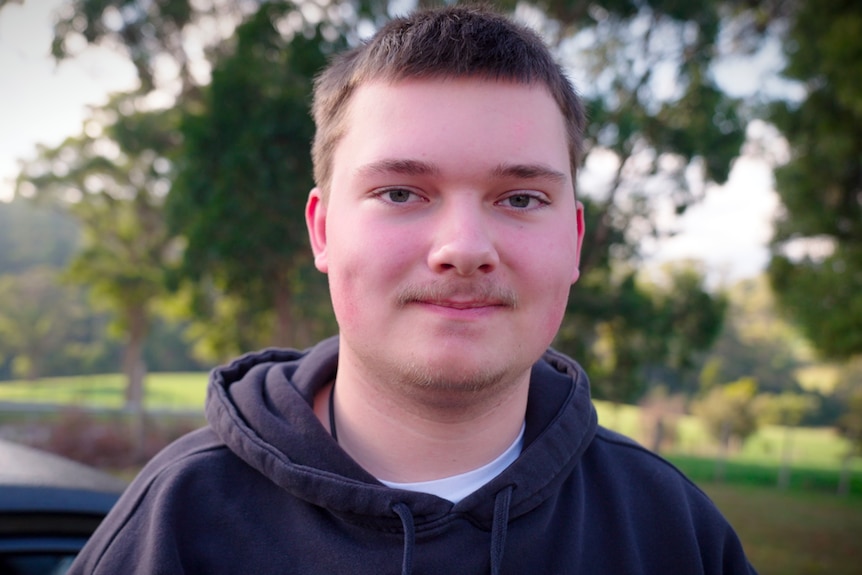 Young man with black hoodie, short hair and a moustache smiles in a park.