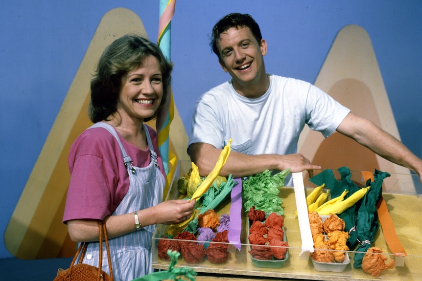 A TV still of Noni Hazlehurst and Simon Burke in C1990, smiling brightly, with a tray of fruit and veggies made of paper.