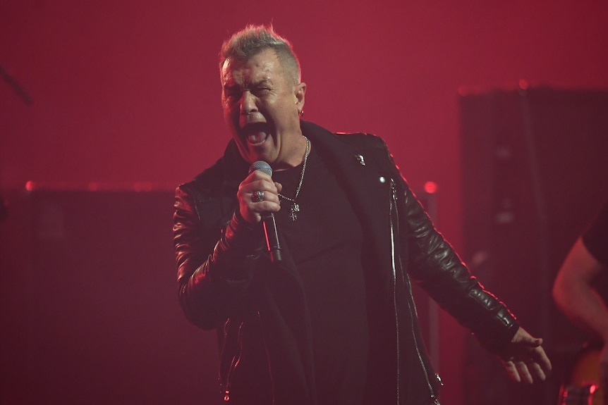 Jimmy Barnes screams into a microphone while wearing a leather jacket onstage.