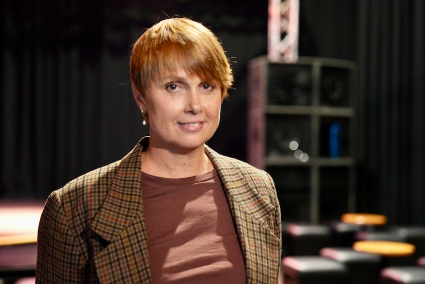 Adele has bright, short hair and wears a checked blazer while smiling for a photo inside a music theatre.