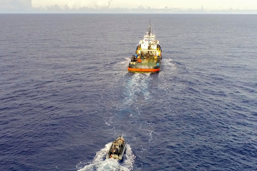 A smaller vessel travels behind a larger vessel carrying large square items at sea.