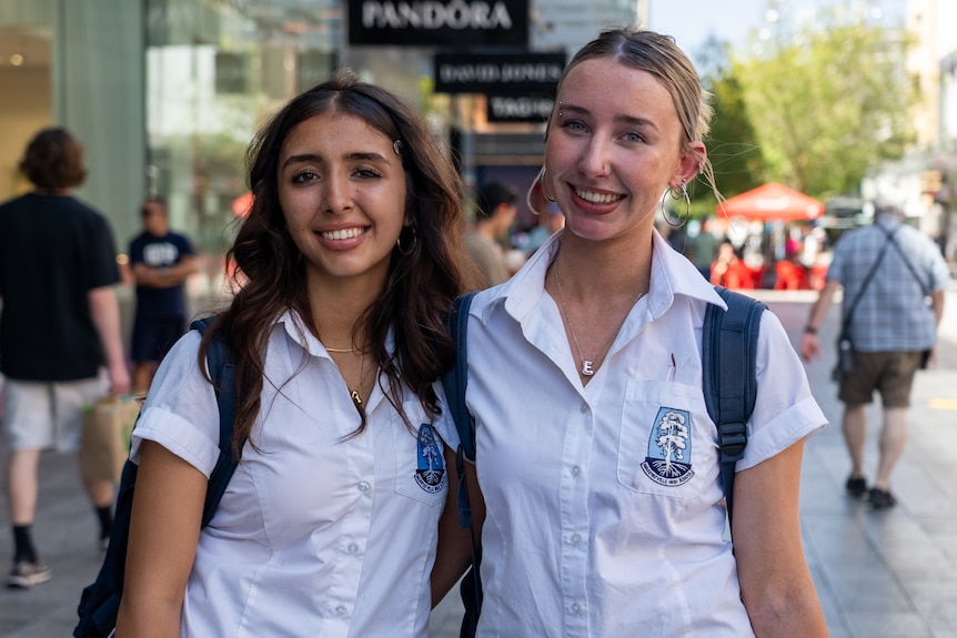Two teenage girls wearing white uniform-style shirts stand together and smile.