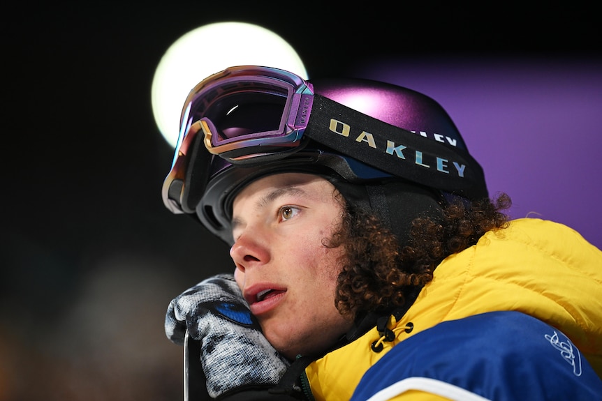 Valentino Guseli wearing helmet and googles rests his head on hand holding snowboard.