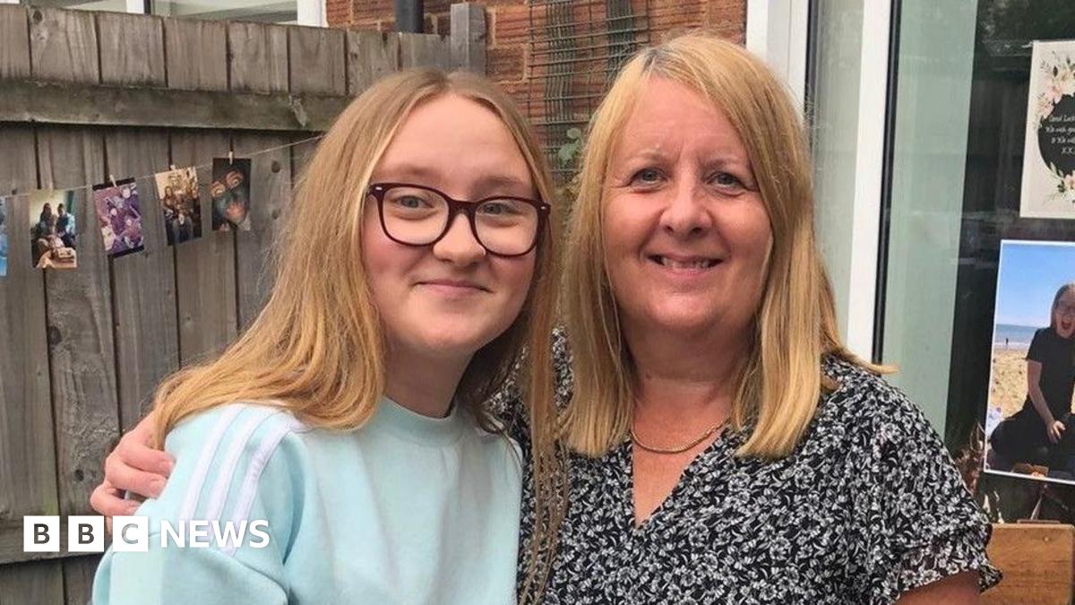 A daughter and mother standing arm in arm in a back garden, with a wooden fence, patio door and various colourful photos in the window and hanging from the fence. The daughter, a young woman, has long strawberry blond hair and wears purple-framed glasses and a light blue jumper with white stripes on the shoulder. The mother has similar hair and wears a black floral blouse, with a thin gold necklace around her neck.