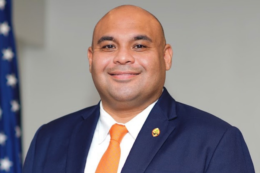 A bald man wearing a blue suit with american flag draped in the background