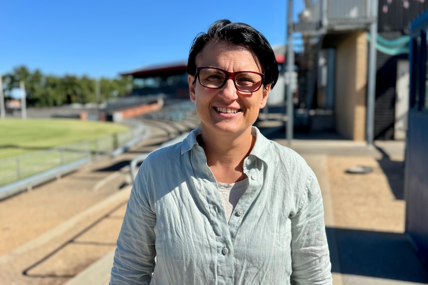 a woman with dark features, short hair smiling at the camera near footy oval 