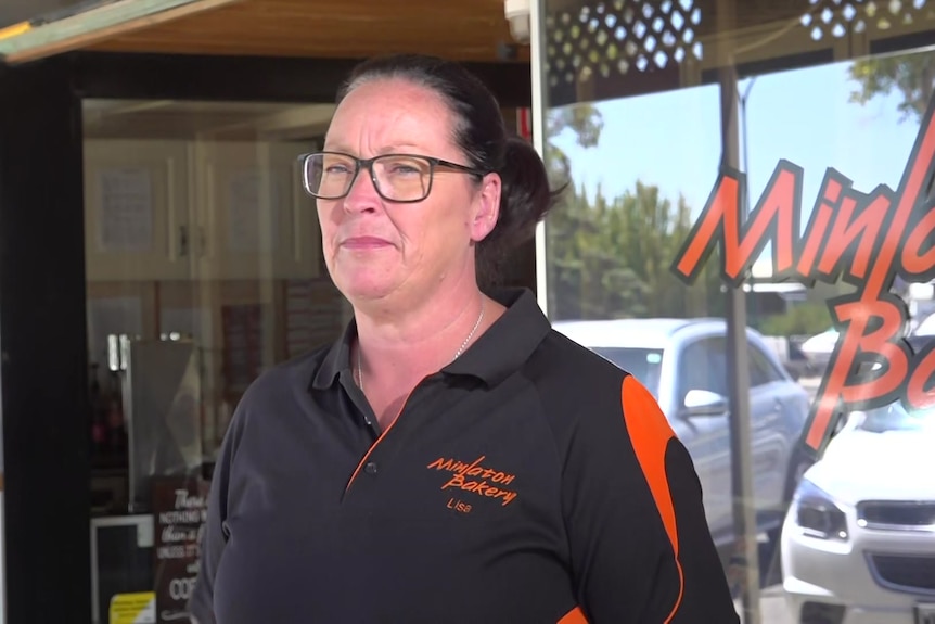 A woman wearing a dark pol-shirt and glasses with her hair tied back stands on the street outside her business