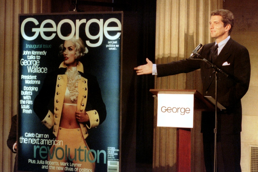 A smiling man in a dark suit stands atr a podium and gestures towards a blown-up magazine cover.
