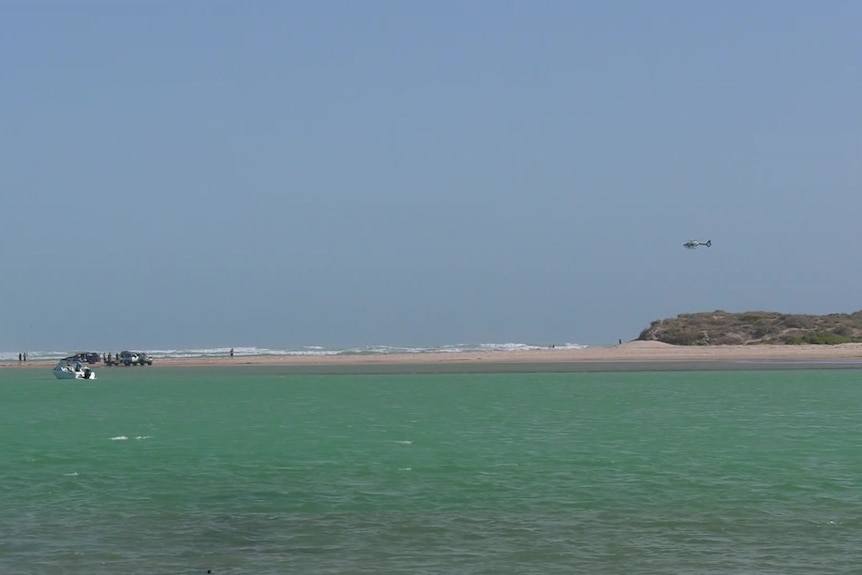 A helicopter flies above land, sand and water
