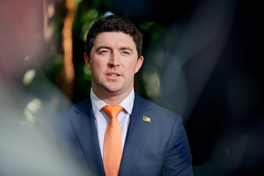 A dark-haired man in a dark suit with a bright tie stands outdoors.
