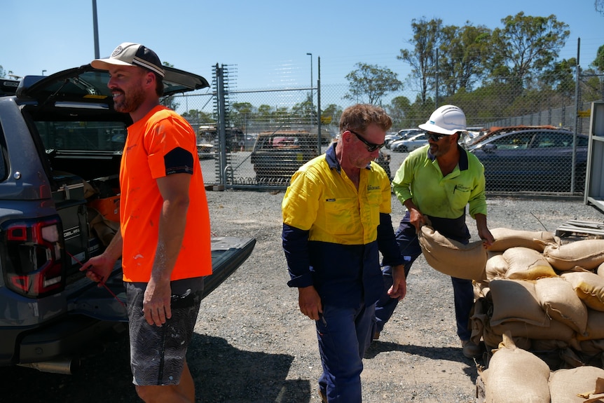 Three men behind a ute wearing high vis throwing sand bags into car.